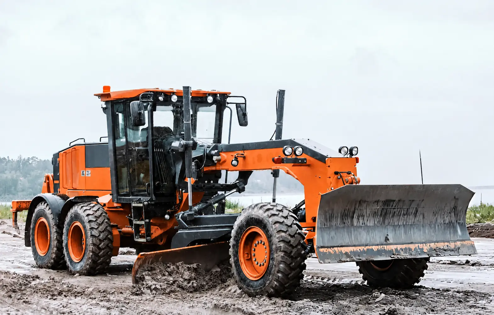 Orange motor grader working on a muddy construction site for road grading, leveling and surface preparation.