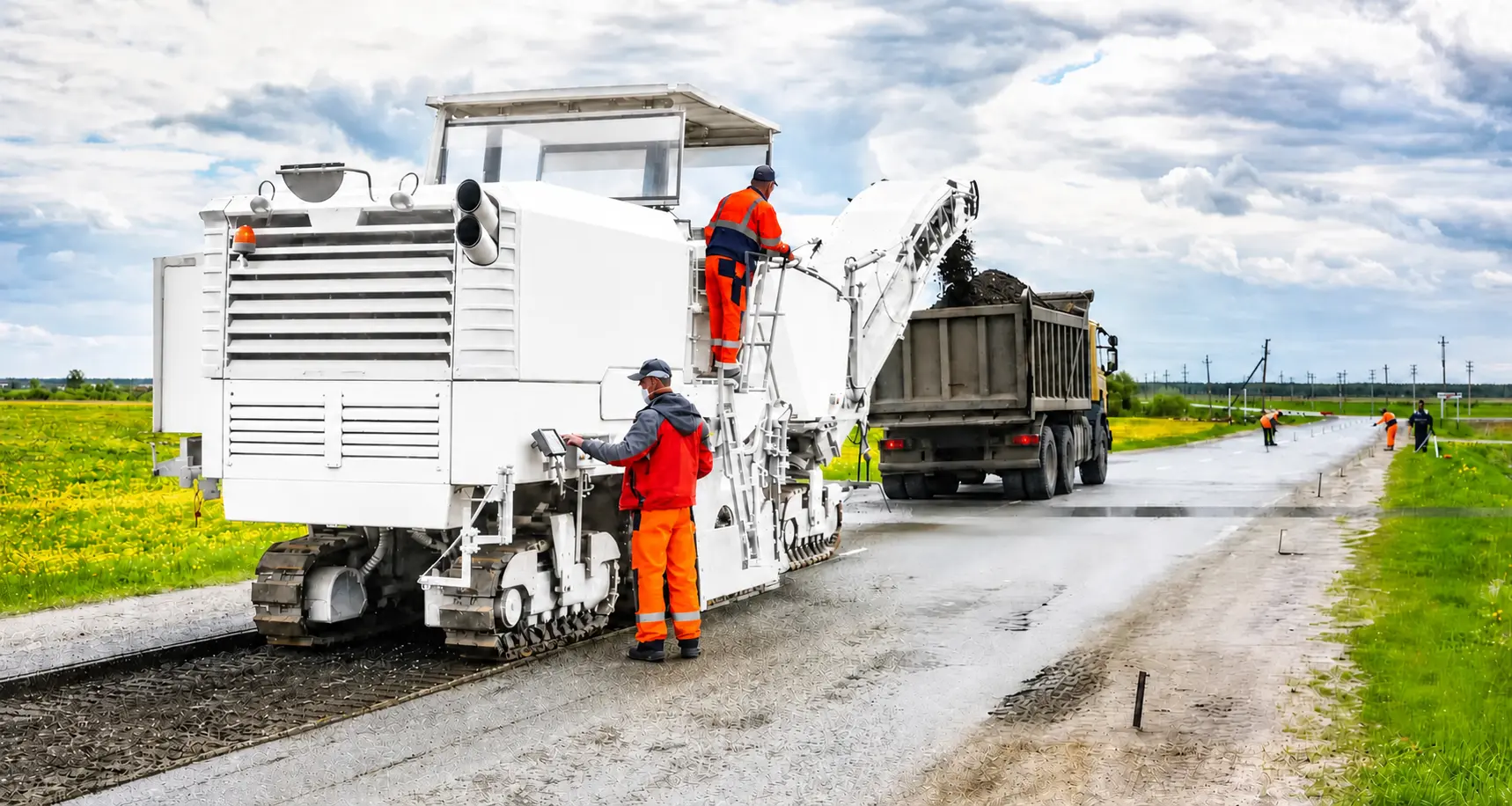 White road milling machine removing asphalt during road construction while workers operate the machine and load milled material into a dump truck.